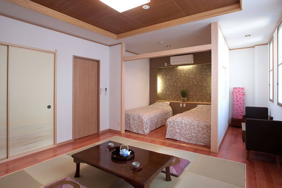 A room with a wooden ceiling, two beds with patterned bedding, a small table with a teapot, and a chair.