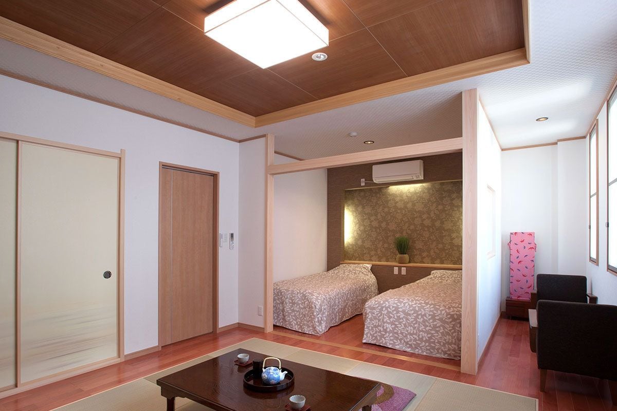 A room with a wooden ceiling, two beds with patterned bedding, a small table with a teapot, and a chair.