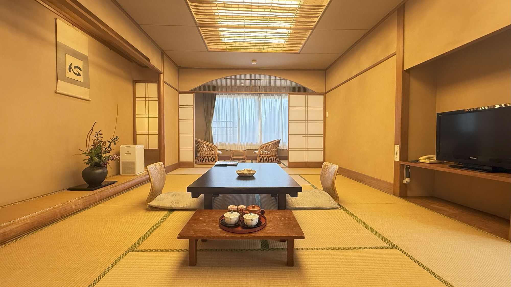 Ein traditioneller japanischer Raum mit Tatami-Bodenbelag, einem niedrigen Tisch mit Sake-Sets, einem Fernseher und einer Schiebetür zu einem Balkon.