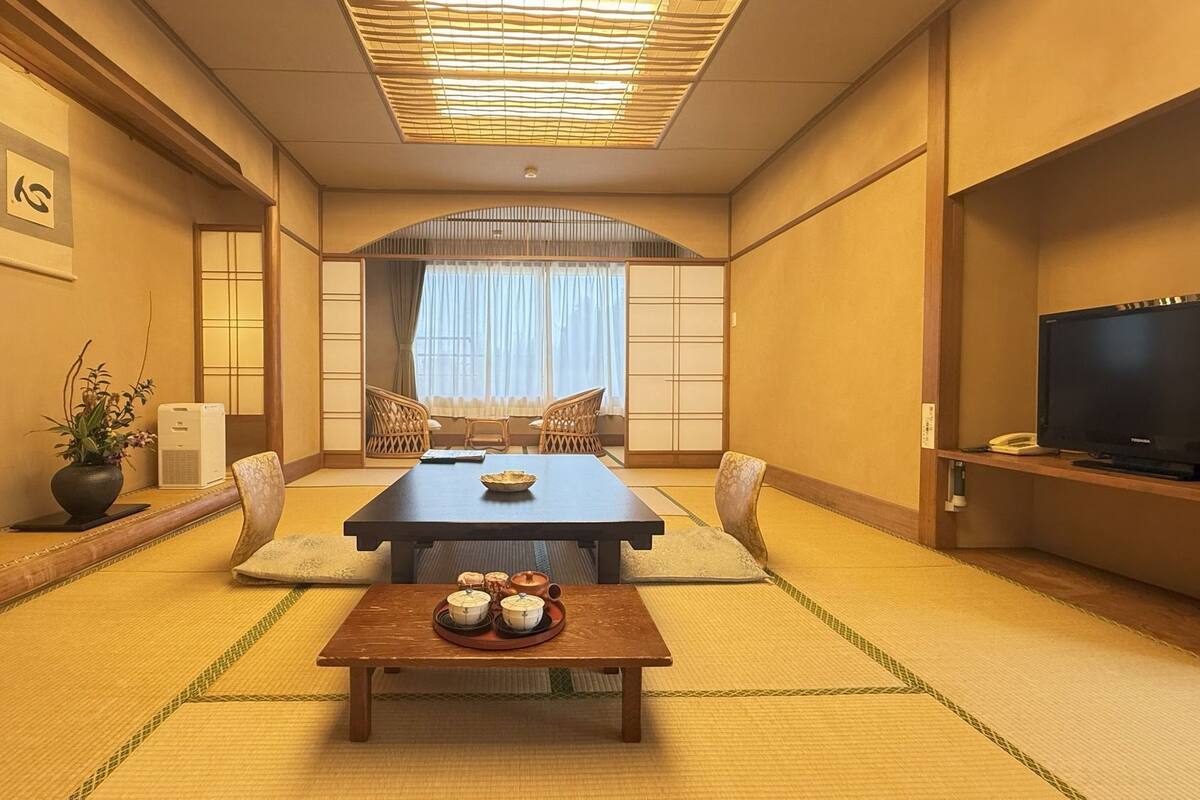 Ein traditioneller japanischer Raum mit Tatami-Bodenbelag, einem niedrigen Tisch mit Sake-Sets, einem Fernseher und einer Schiebetür zu einem Balkon.