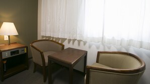 Ein Hotelzimmer mit Schreibtisch, zwei Sesseln, einer Lampe und einem Fenster mit leichtem Vorhang.