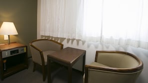 Habitación de hotel con escritorio, dos sillones, una lámpara y una ventana con cortinas translúcidas.
