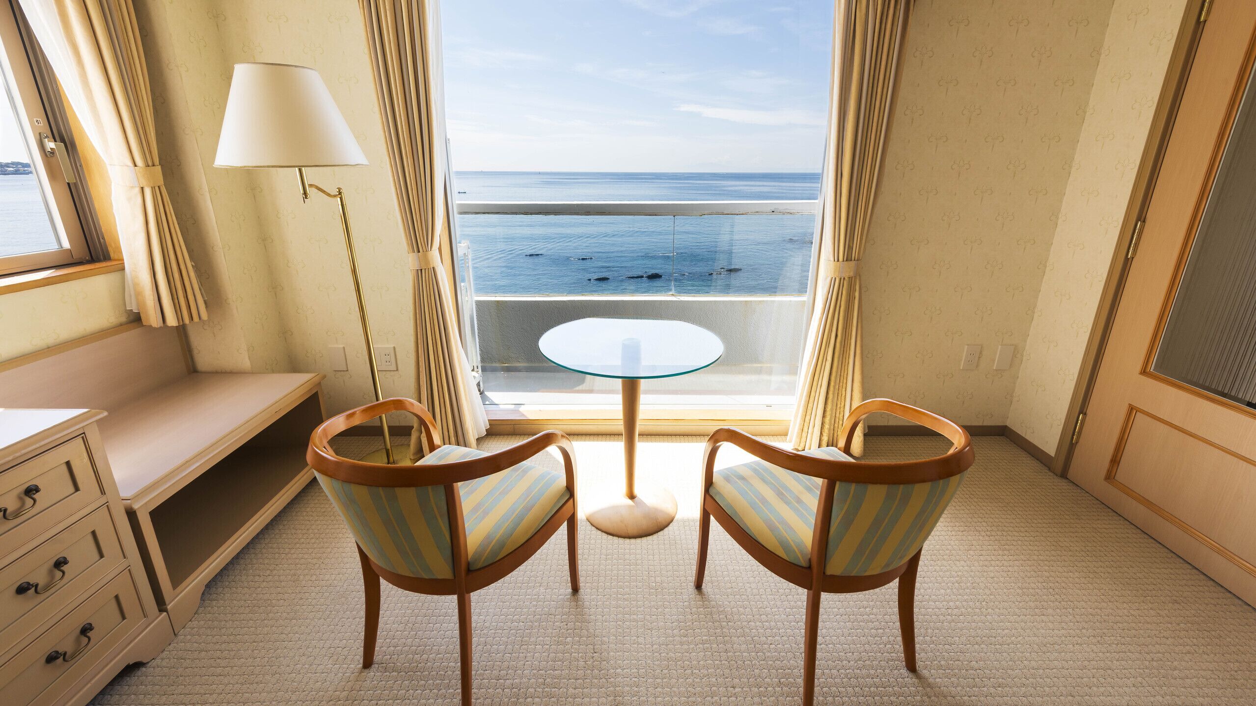 A room with a view of the sea, a round glass table, two striped chairs, a lamp, and a wooden dresser.