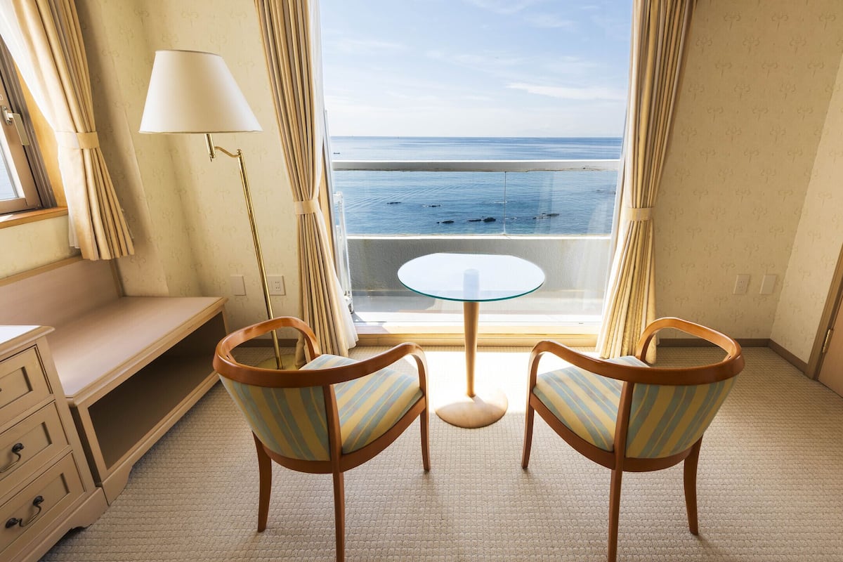 A room with a view of the sea, a round glass table, two striped chairs, a lamp, and a wooden dresser.