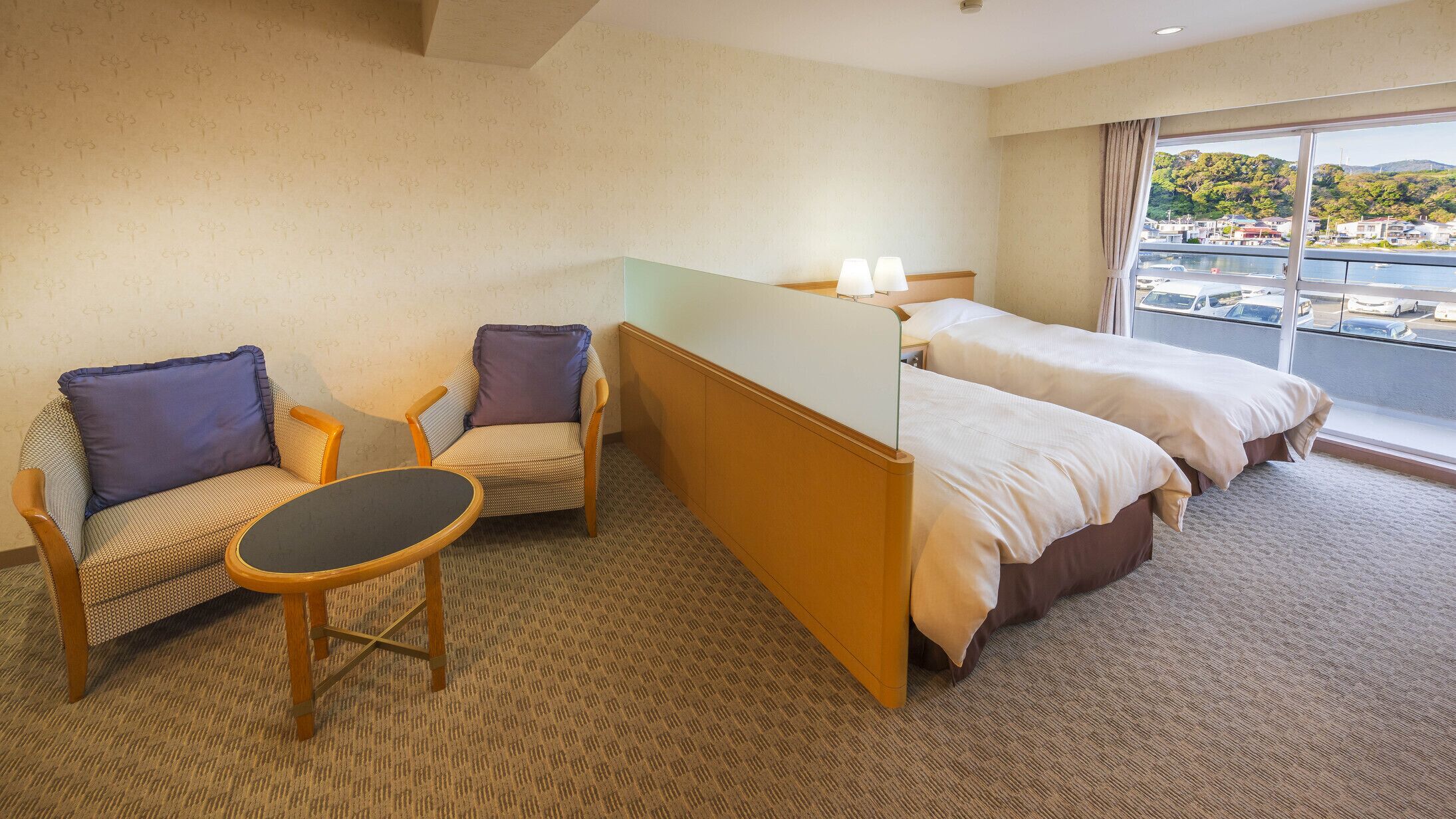 A hotel room with two beds, a sofa, a small table, and a view of a body of water and hills.