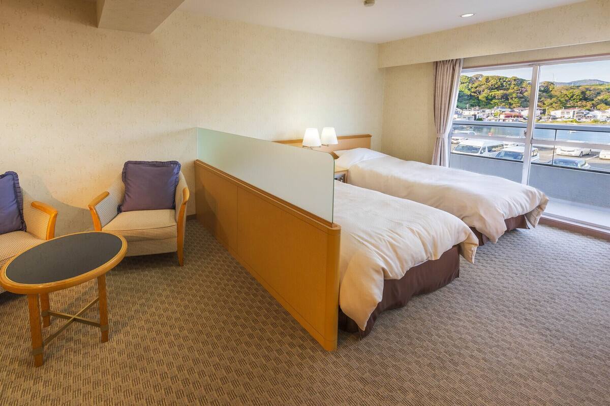 A hotel room with two beds, a sofa, a small table, and a view of a body of water and hills.