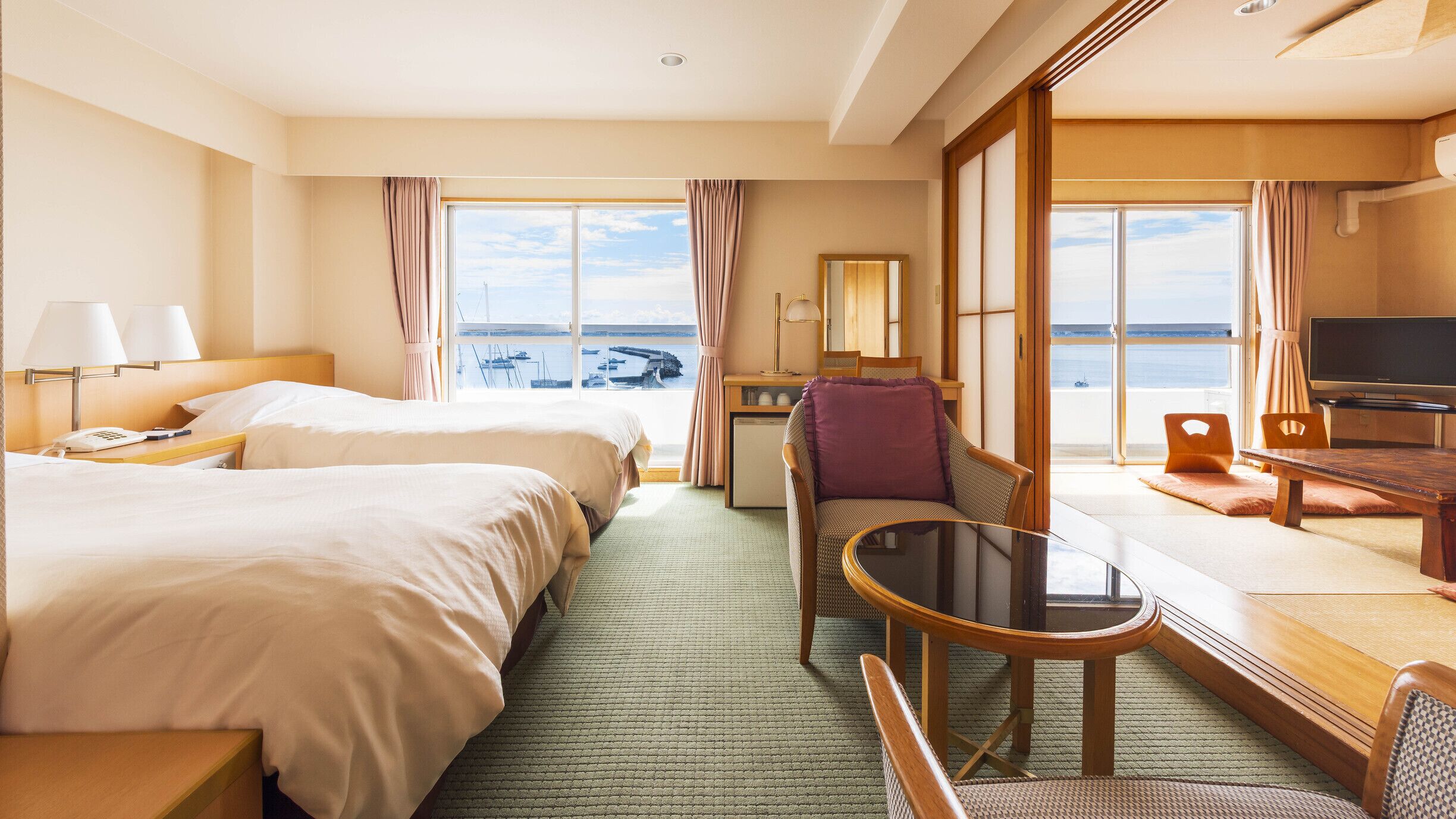 Een hotelkamer met twee bedden, een bureau, een stoel, een televisie en uitzicht op zee.