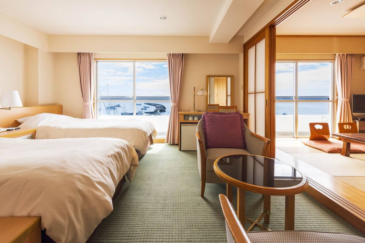 A hotel room with two beds, a desk, a chair, a TV, and a view of the sea.