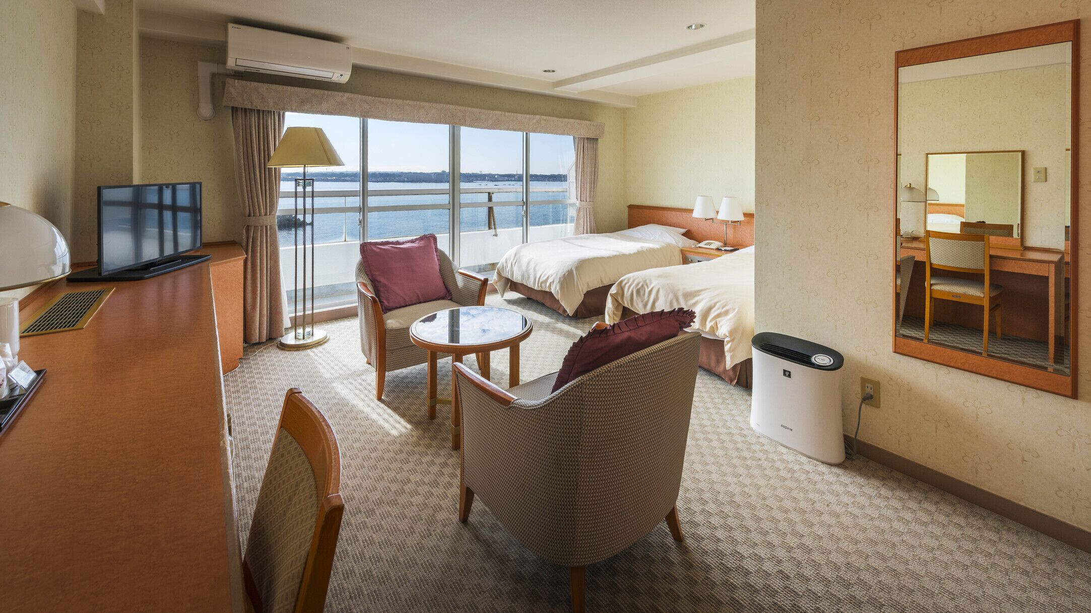 A hotel room with a bed, a desk, a chair, a small table, a TV, and a window with a view of the sea.