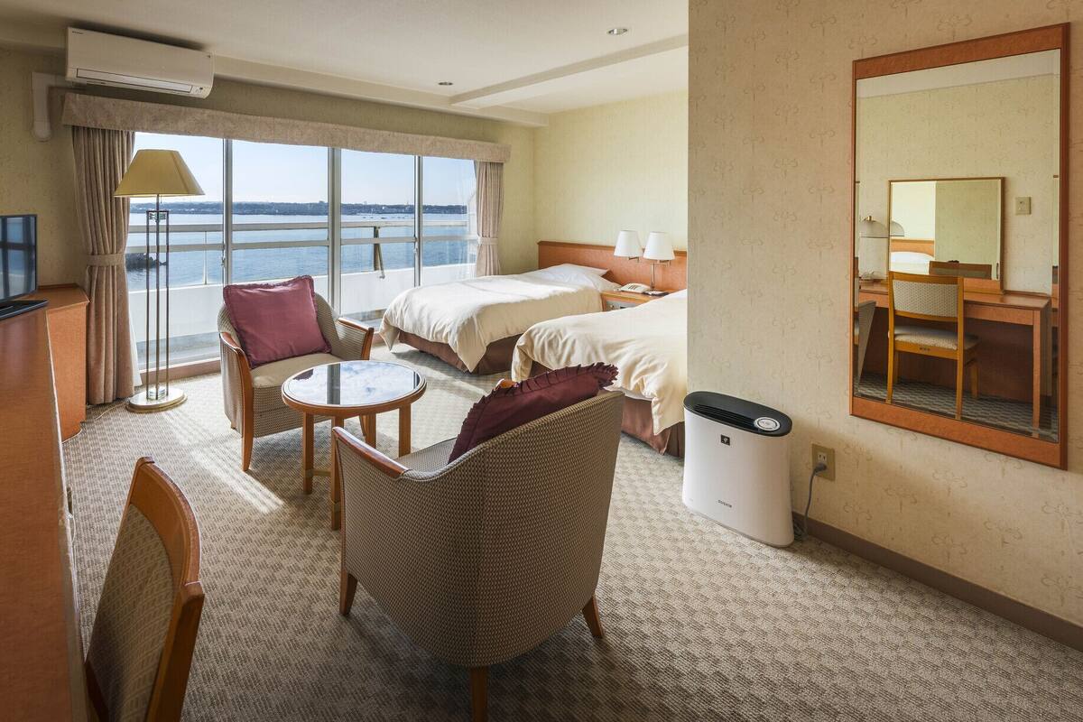 A hotel room with a bed, a desk, a chair, a small table, a TV, and a window with a view of the sea.