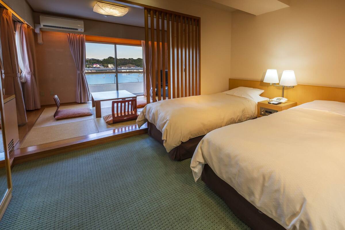 A hotel room with two beds, a small table, a chair, a view of a body of water, and a window with curtains.