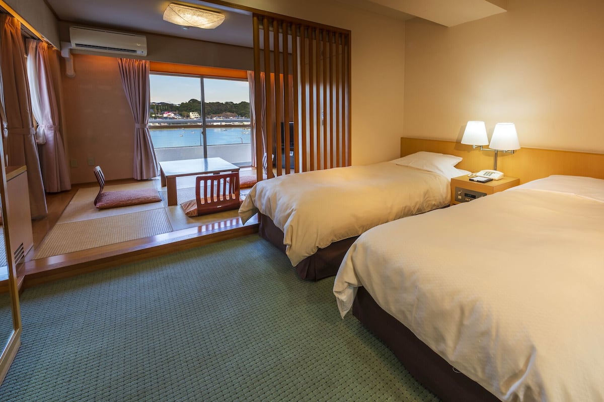 Hotelkamer met twee bedden, een kleine tafel, een stoel en uitzicht op water, met gordijnen voor het raam.