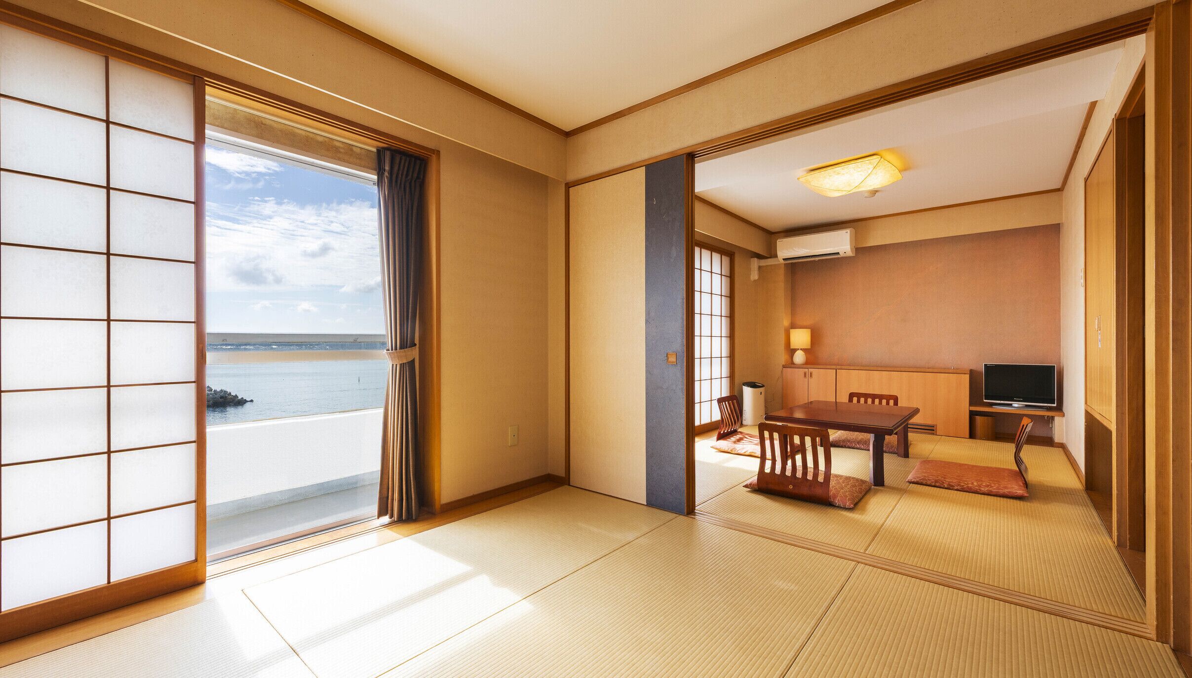 A traditional Japanese room with tatami flooring, a sliding door to a balcony, a small table with chairs, and a television.