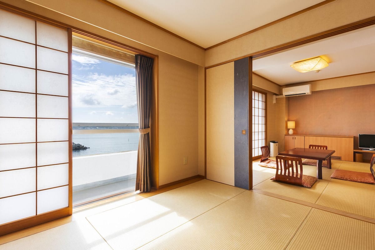 A traditional Japanese room with tatami flooring, a sliding door to a balcony, a small table with chairs, and a television.