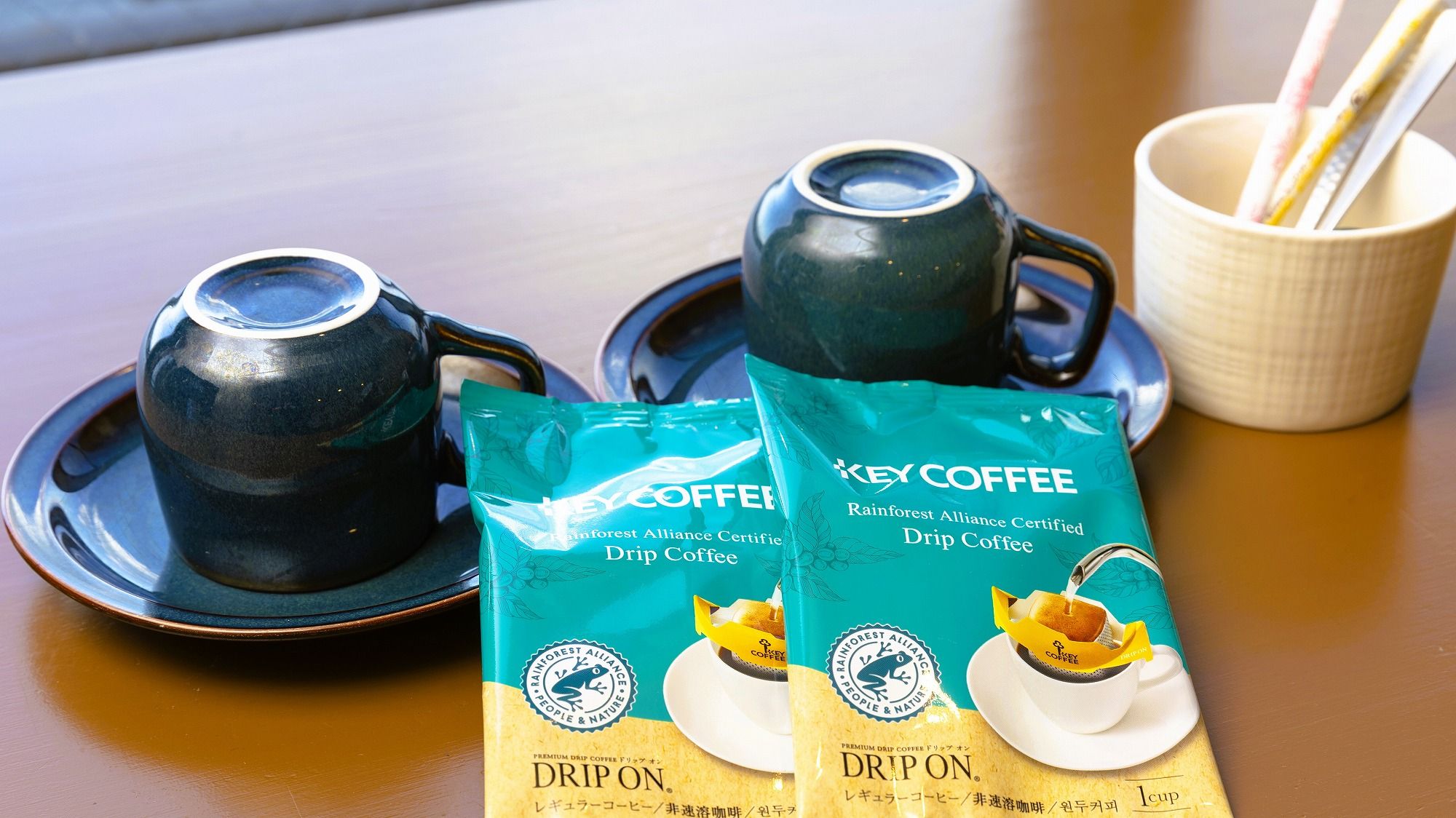 To kopper sort kaffe på underkopper, en hvid kop med sugerør, og to pakker Key Coffee Rainforest Alliance Certificeret Drip Kaffe.