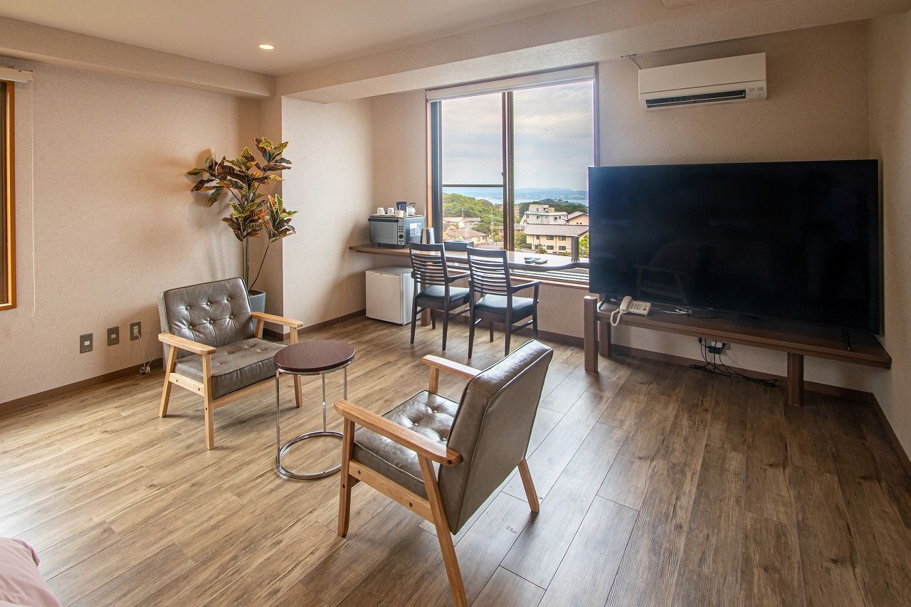 Una sala de estar moderna con un televisor grande, dos sillones de cuero, una mesita y vistas a la ciudad a través de amplios ventanales.
