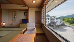 A traditional Japanese room with tatami flooring, a low wooden table, a sofa, a television, and a view of a modern building and greenery outside. - Noji Onsen Hotel (Fukushima)