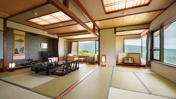 A traditional Japanese-style room with tatami flooring, a low table with cushions, a television, and a large window offering a view of the mountains. - Noji Onsen Hotel (Fukushima)
