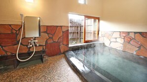 Una bañera de hidromasaje con paredes de piedra y una ventana, una zona de ducha con espejo, y suelo de piedra.