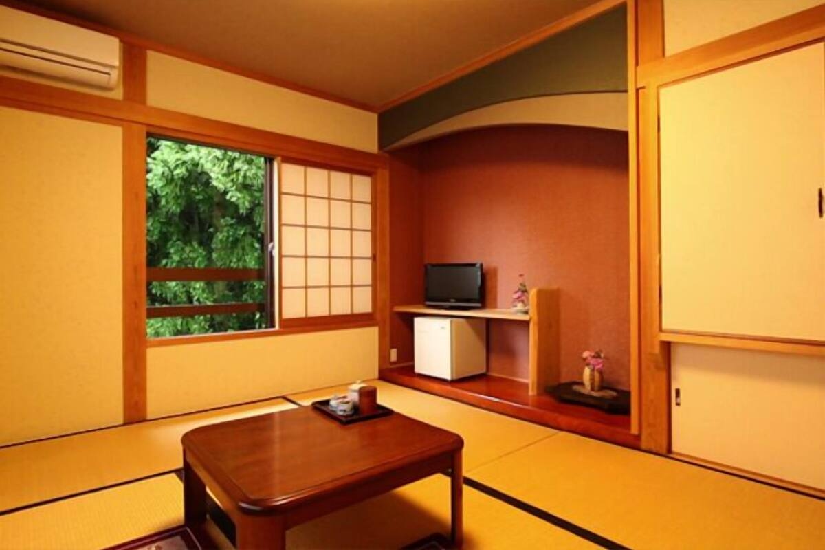 A traditional Japanese room with tatami flooring, a low table, a television on a shelf, and a view of greenery outside.
