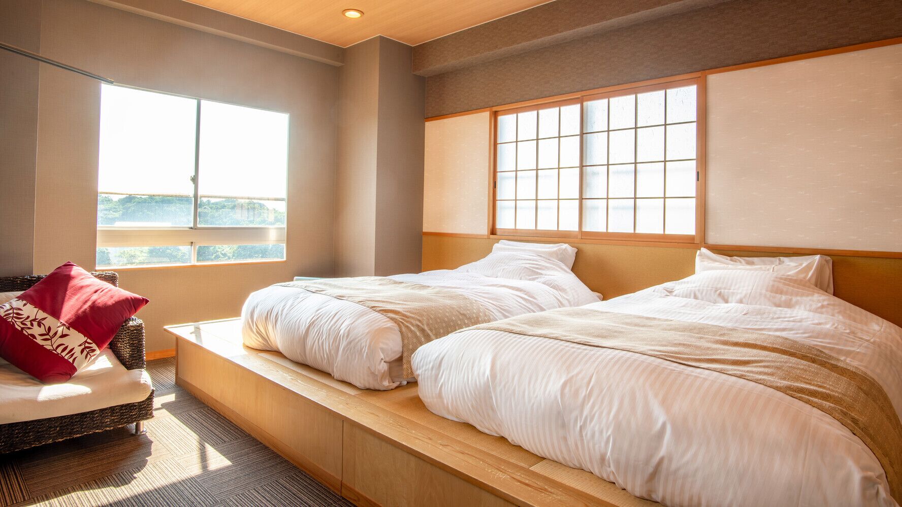 Ein Hotelzimmer mit zwei Betten, Blick ins Freie und einer Sitzecke mit einem roten Kissen.
