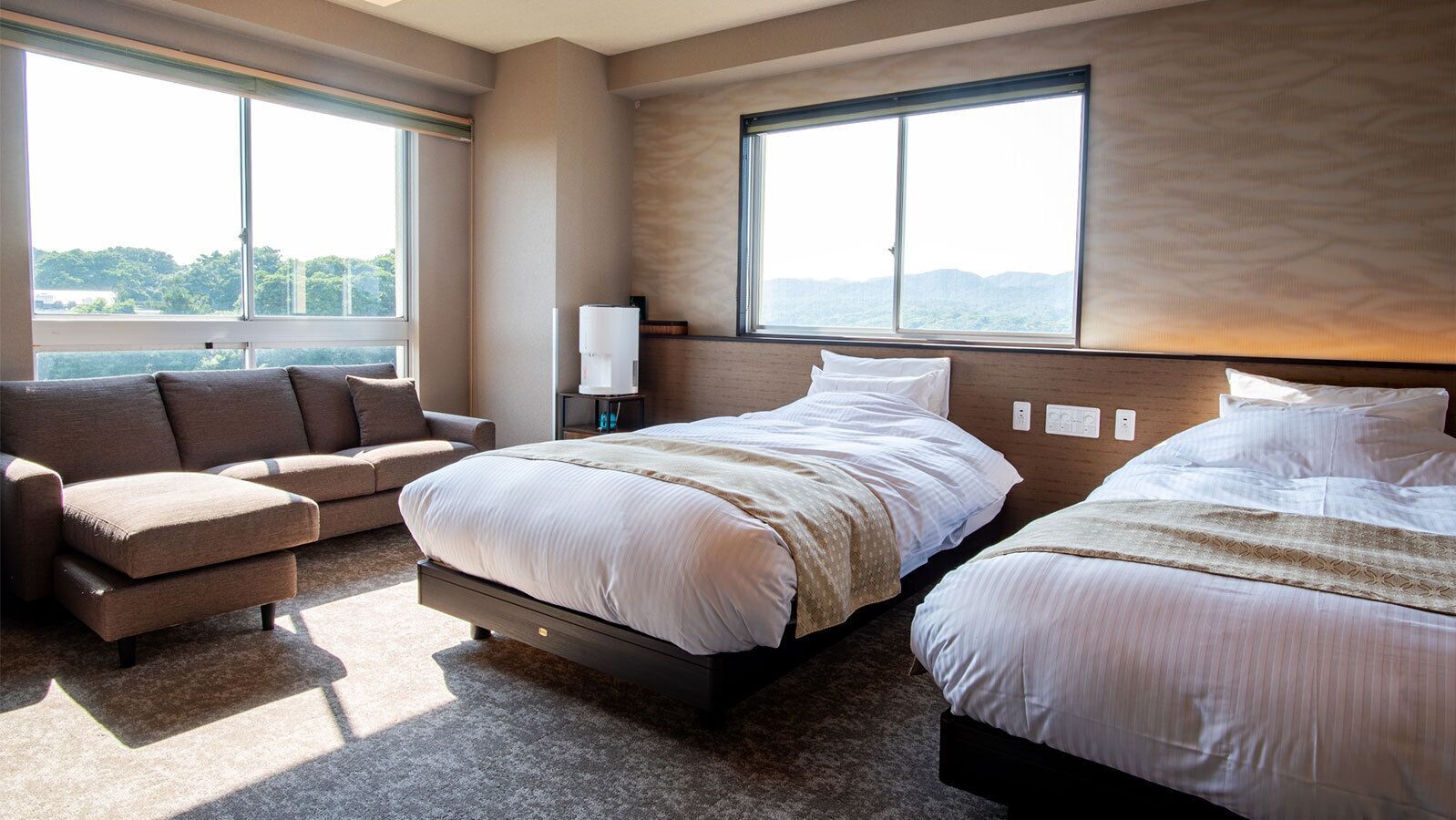 Habitación de hotel con dos camas, un sofá, una mesita de noche con lámpara y ventanas amplias con vistas.