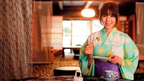 Una persona vestida con traje tradicional sostiene una tetera y un plato, con una taza de té sobre la mesa.