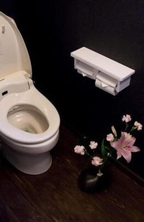 Des toilettes avec un abattant et un couvercle blancs, un porte-rouleau de papier toilette blanc fixé au mur, et un vase avec des fleurs roses posé sur le sol.