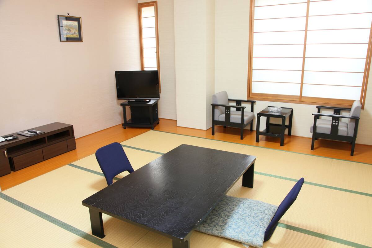 Ein Zimmer mit Tatami-Böden, einem schwarzen Tisch, blauen Stühlen, einem Fernseher und einem kleinen Schrank.
