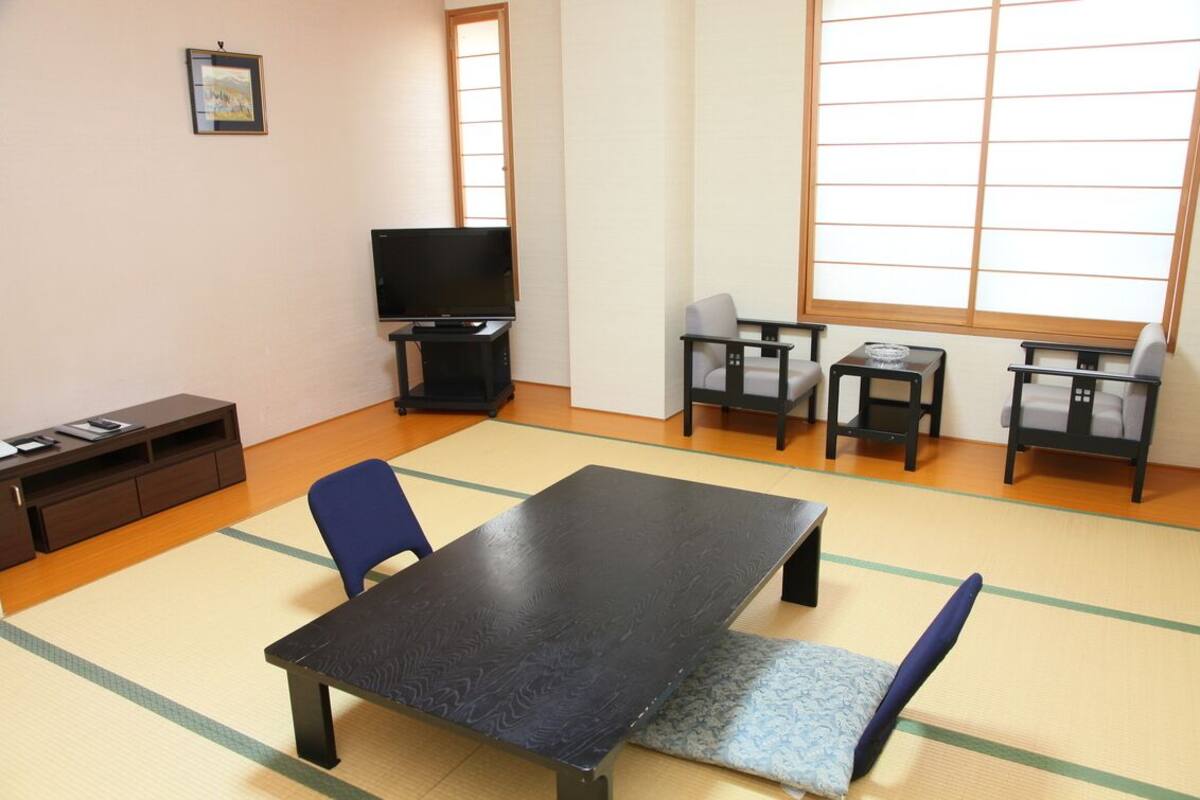 Ein Zimmer mit Tatami-Boden, einem Tatami-Tisch, zwei Stühlen, einem Fernseher und einem kleinen Schrank.