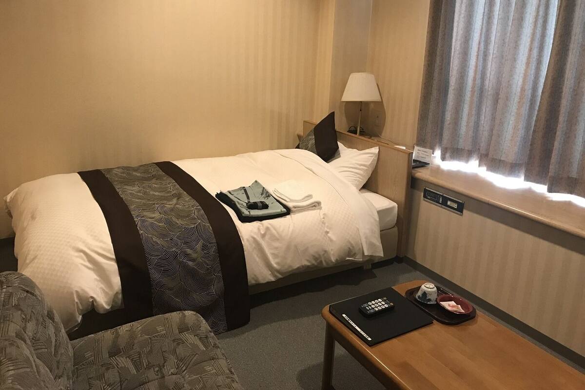 Ein Hotelzimmer mit Bett, Schreibtisch, Stuhl, Lampe und einem fensterbepflanzten Fenster.