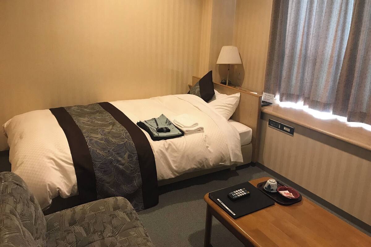 Ein Hotelzimmer mit einem Bett, einem Nachttisch mit Fernbedienung, einer Lampe und einem Fenster mit Vorhängen.