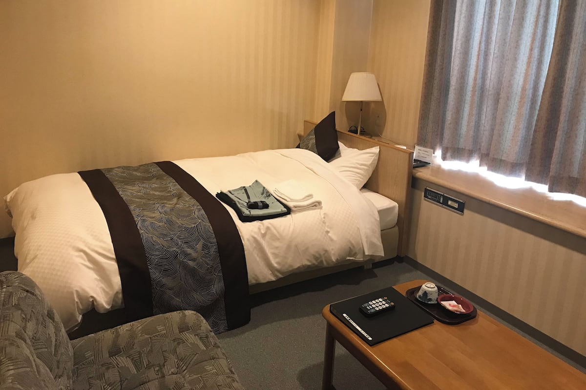 Habitación de hotel con cama, mesita de noche con control remoto, lámpara y ventana con cortinas.