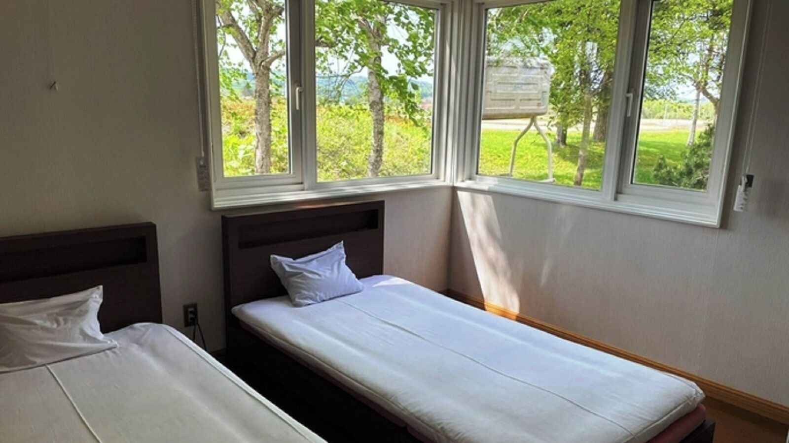 Un lit simple avec une literie blanche et une tête de lit en bois, dans une pièce dotée d’une grande fenêtre donnant sur des arbres et une zone herbeuse.