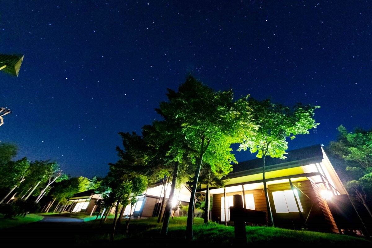 A modern house with a large window, surrounded by trees and a clear night sky.