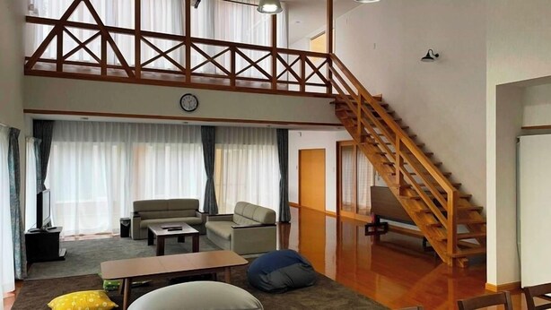 A spacious loft-style room with a wooden staircase, a sofa, a coffee table, and a large window with curtains. - Cottage Villa Top (Shintotsukawa)
