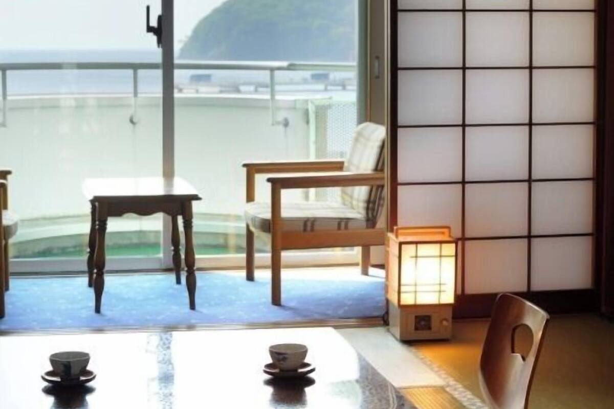 Um quarto com porta de vidro deslizante, uma mesa de madeira com duas cadeiras, uma mesa pequena com uma xícara e um tradicional biombos japonês shoji.