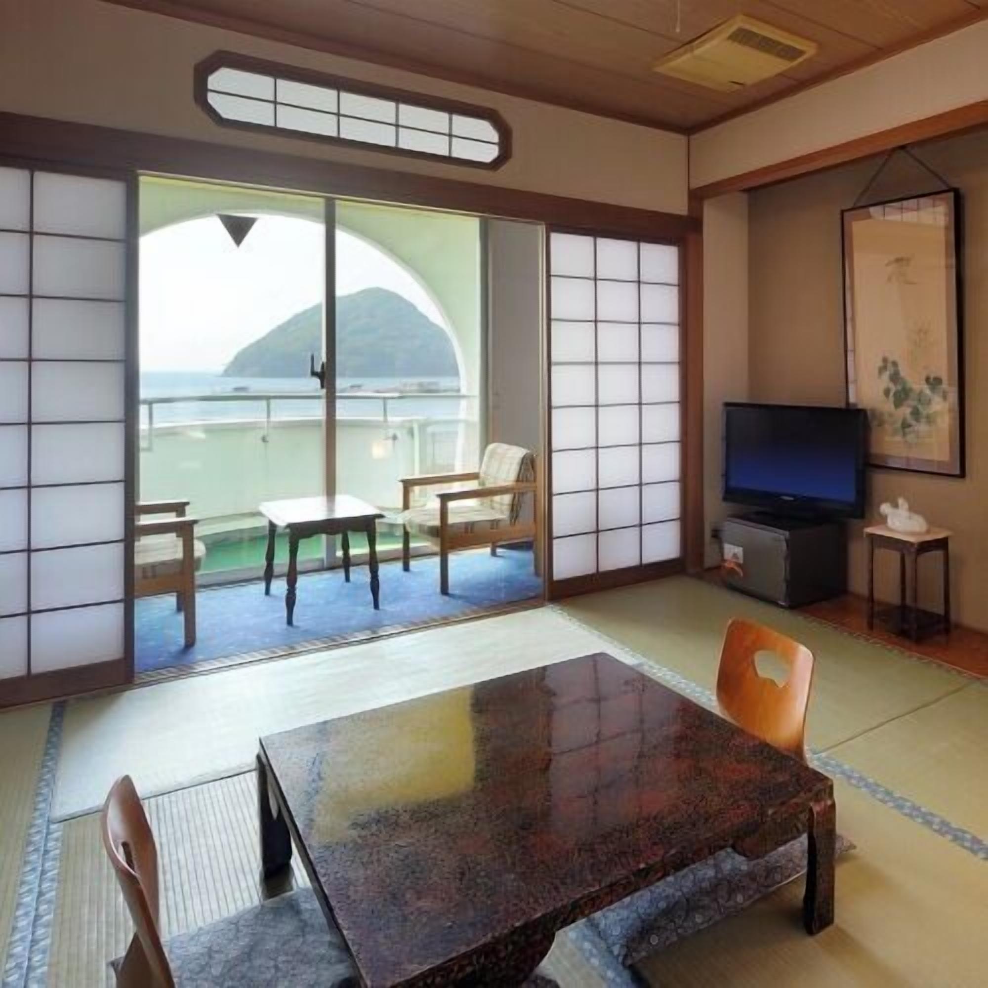 Una habitación japonesa tradicional con suelo de tatami, una puerta corredera de vidrio a un balcón, una mesita baja y un televisor.