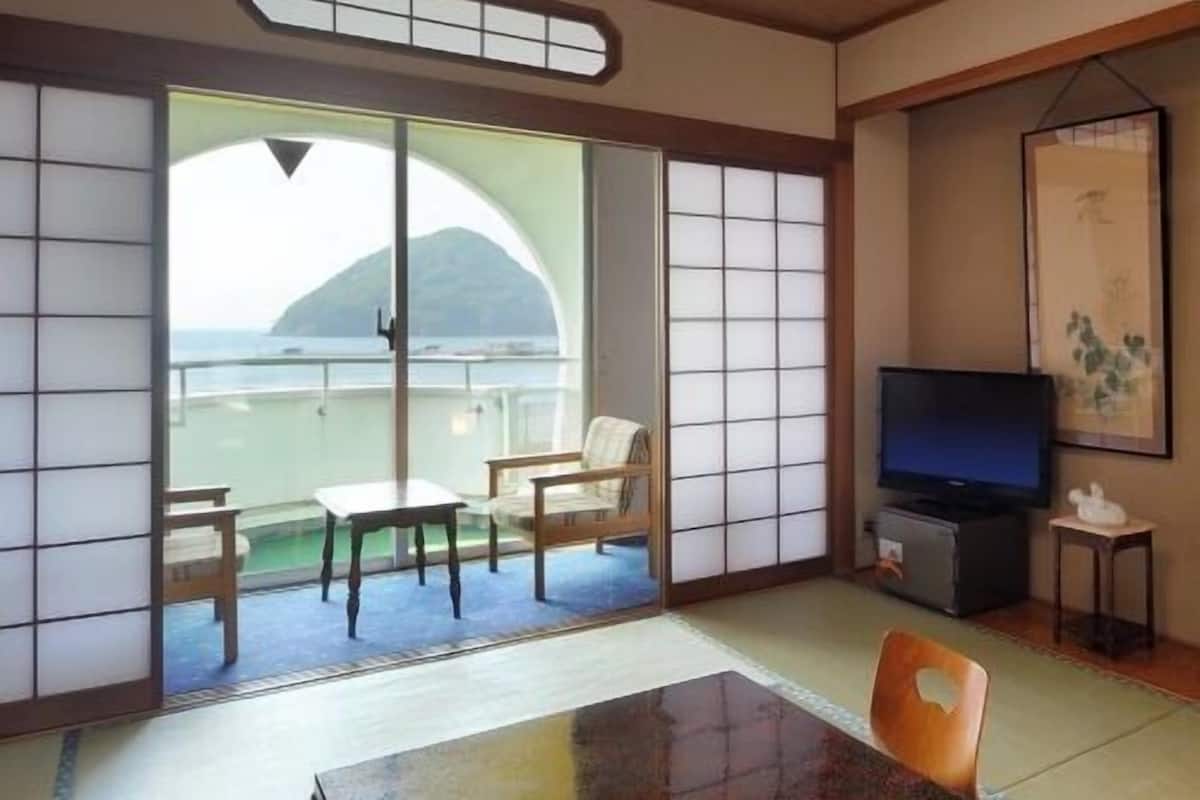 Um quarto japonês tradicional com tatames, uma porta de vidro deslizante para uma varanda, uma mesa baixa e uma televisão.