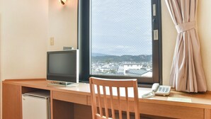 A hotel room with a wooden desk, a computer monitor, a telephone, and a window with a view of buildings and mountains. - Hotel Primrose Saito (Saito)