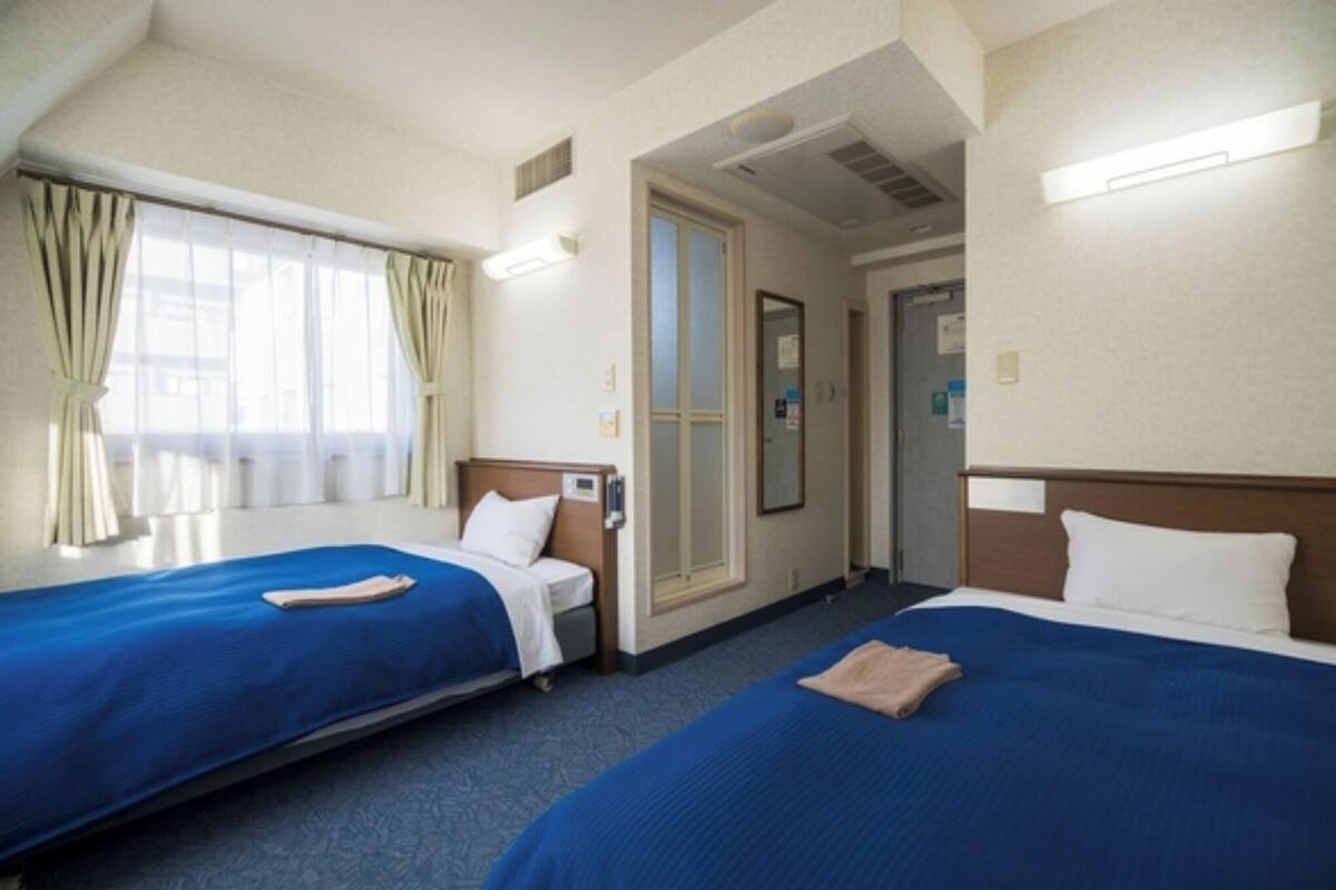 A hotel room with two beds, a window with curtains, a door, and a wall-mounted light fixture.