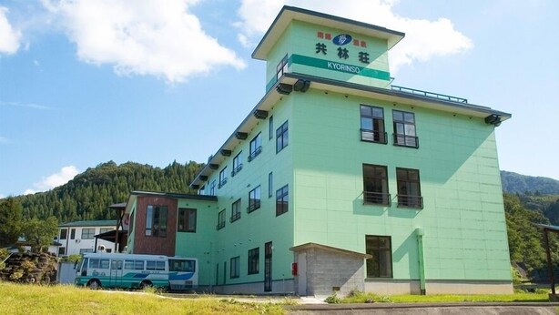 A multi-story building with a green facade, a bus parked in front, and a mountainous backdrop. - Kyorinso (Yokote)