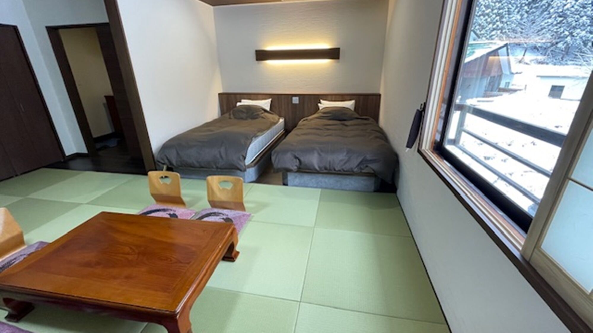 A room with two beds, a wooden table, and a window with a view of a snowy landscape.
