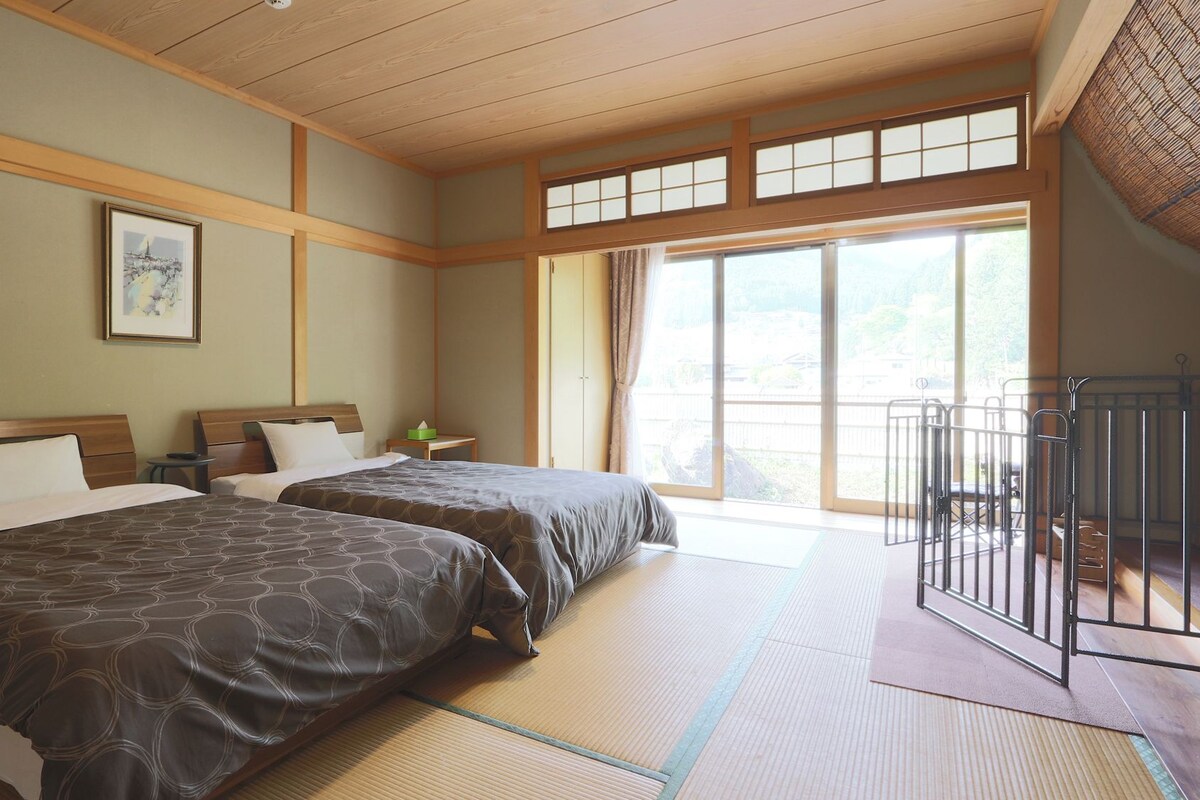 A hotel room with two beds, a large window, tatami flooring, and a view of the outdoors.