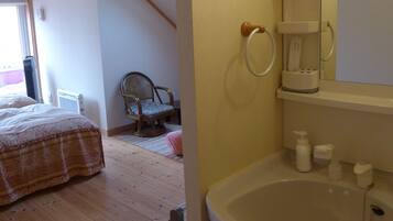 A bedroom with a bed, a chair, a window, and a bathroom with a sink and mirror.