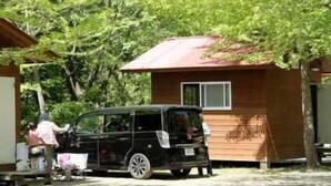 A small wooden cabin with a red roof, a black vehicle parked in front, and a person standing near the entrance. - Hotel Silk Villa (Kimitsu)