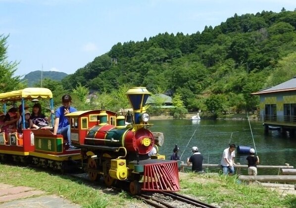 A small, colorful train on tracks with passengers, a body of water, and greenery in the background.