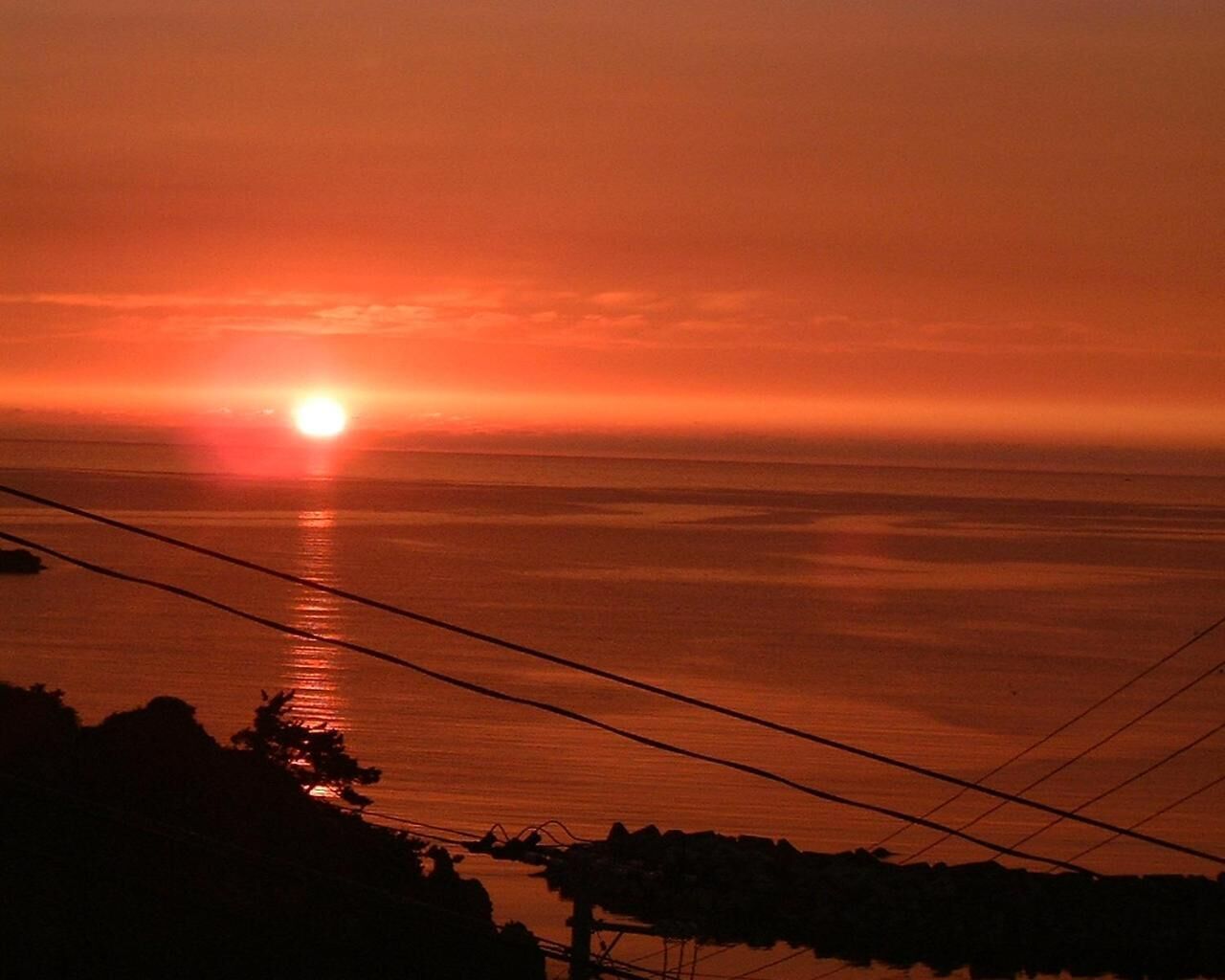 A sunset over a calm sea with power lines in the foreground.