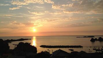 A coastal sunset with the sun low on the horizon, reflecting on the water. There are rocky outcrops and a breakwater visible.
