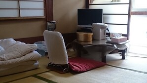 A traditional Japanese room with tatami flooring, a futon bed, a low table with a television, and a window with shoji screens. - Komecho Ryokan (Imabari)
